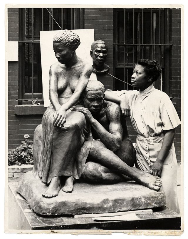 A black and white photograph of the artist Augusta Savage sitting by her sculptures.