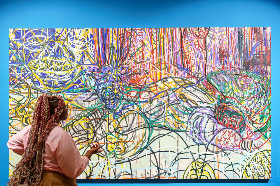 A woman with long braids is looking at a vibrant abstract painting, which hangs on a bright blue wall.