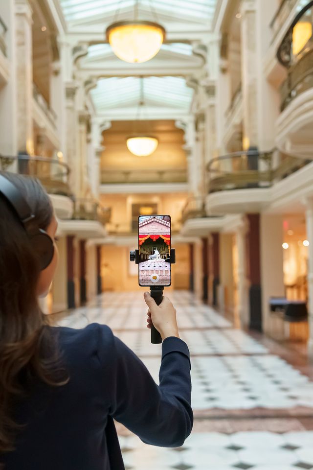 A person wearing headphones stands in a historic building. She is holding a phone up and looking at its screen.