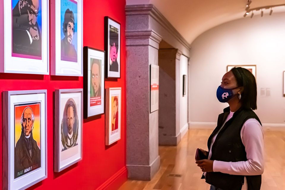 A woman stands in front of a red wall showing 7 graphic artworks 