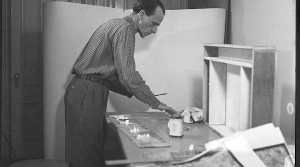 Artist Joseph Cornell standing at worktable with materials for his artworks around him