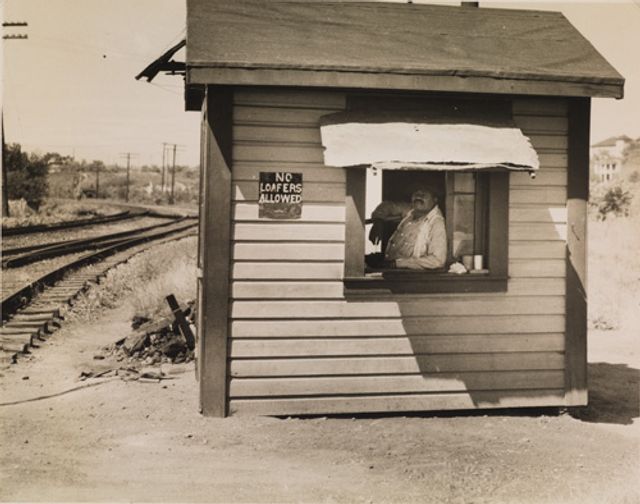 McNeill's gelatin silver print of a man in a small house by the railroad.