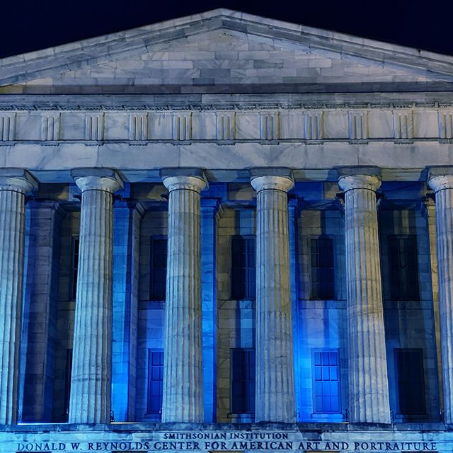 A photograph of the facade of the Smithsonian American Art Museum at night with blue lights honoring healthcare workers.