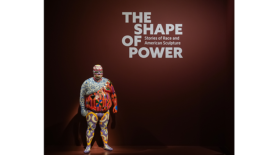 Sculpture of a person completely covered with multiple colorful, intricate patterns standing against a dark red wall with the exhibition title "The Shape of Power: Stories of Race and American Sculpture."