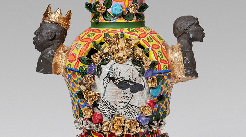 A detailed crop of a highly textured ceramic vessel. The center shows a painting of the rapper Notorious B.I.G. The side handles are busts of Notorious B.I.G. on one and Tupac Shakur on the other.