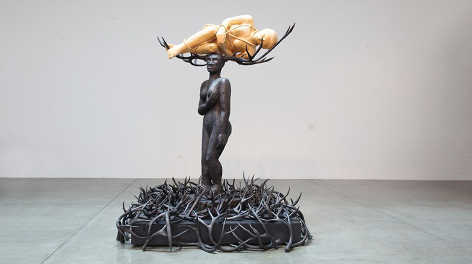 Sculpture of a woman. Massive antlers on her head cradle a delicate, translucent adult figure in a fetal position, like a creature preparing to emerge from its cocoon. Scattered on the ground below are antlers.