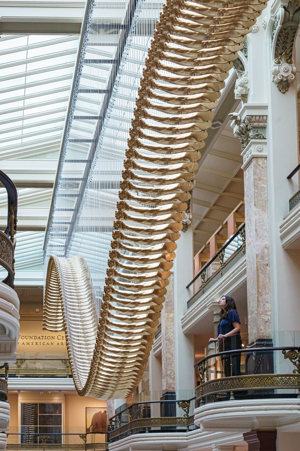 A person standing on a balcony in a historic museum looks at an undulating artwork made up of 200 golden-cast arms hanging from the ceiling.