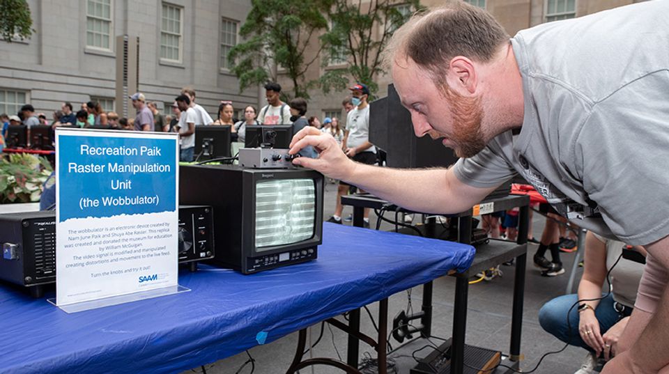 A man turns the nobs of a box sitting atop a small black and white tv monitor.