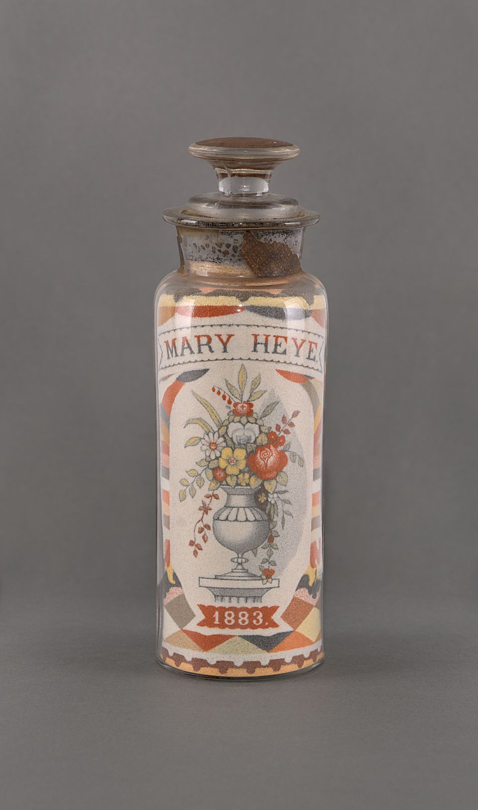 A tall, slim bottle holding sand that says Mary Heye and has an ornate design.