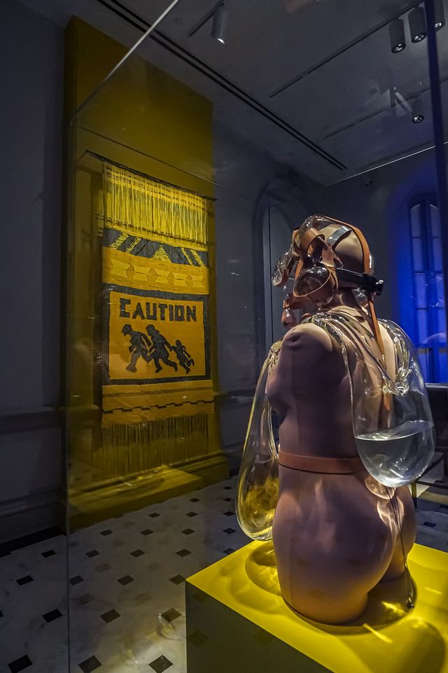 Two artworks in a gallery. On the back wall, a yellow woven tapestry that says "Caution" hangs on the wall. In the foreground is a glass sculpture in the shape of a gas mask and backpack on a mannequin.