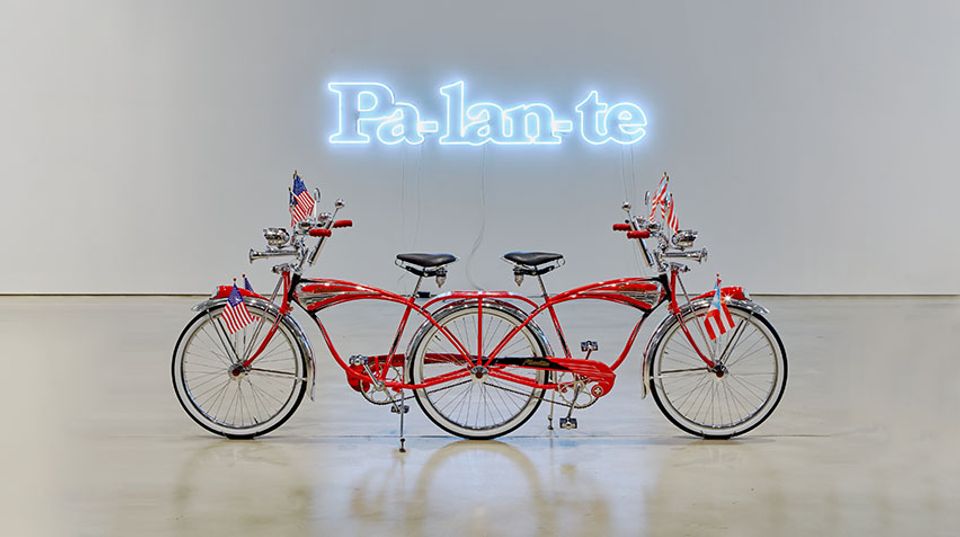 Bicycle sculpture with a neon artwork above it that reads "Pa-lan-te."