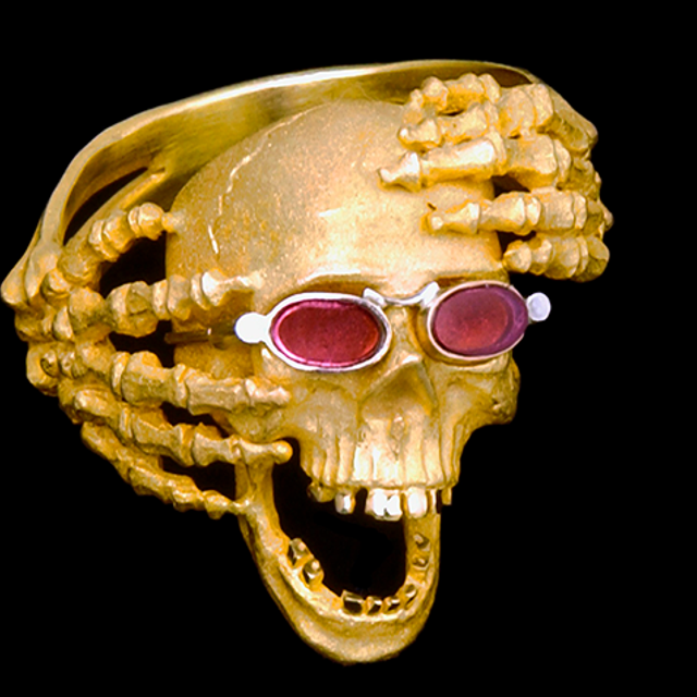 Sculpture that shows gold skeleton hands reaching around a gold skull that is wearing rose-colored glasses.