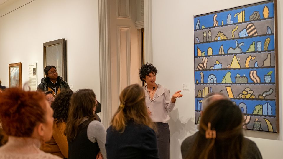 A person stands in front of an abstract painting. She is speaking to a group of onlookers.