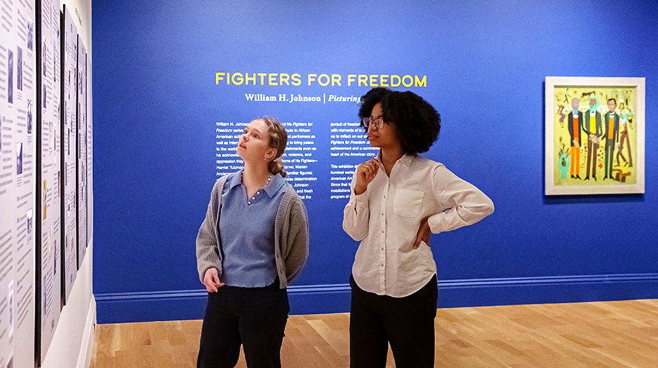 Two visitors look at a timeline on a wall. Behind them is a painting hanging on a blue wall and the words "Fighters for Freedom" with text underneath.