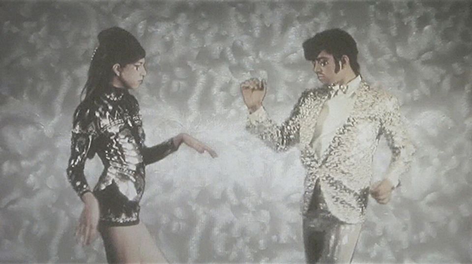 Two people in 60s mod clothing face each other. They stand in a gray, gauzy space.