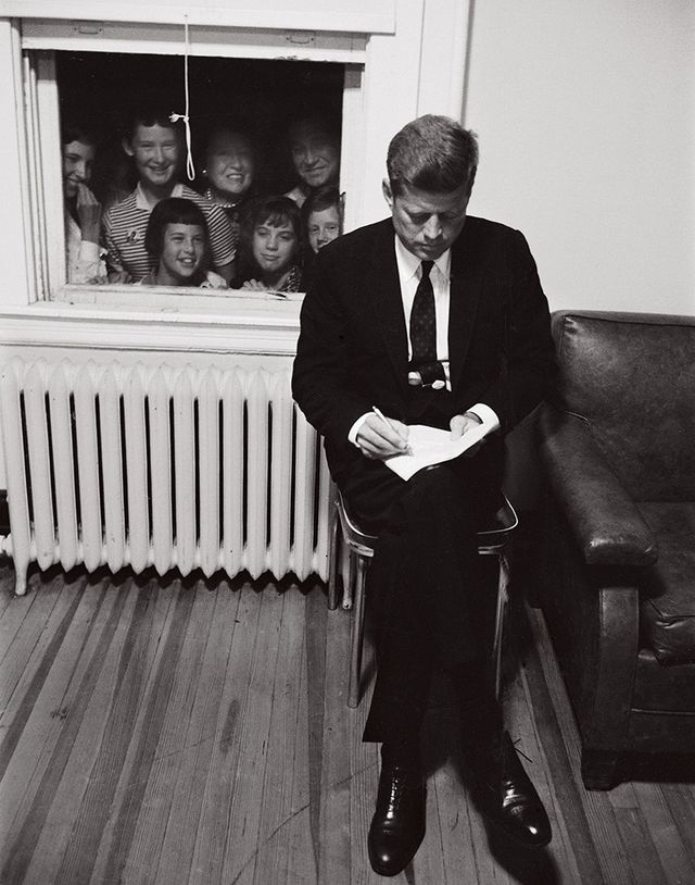 Seated Kennedy looking at notes while a crowd peers in through a window behind him