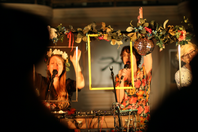 Two performers sing into microphones. Colorful wooden frames and flowers hang in front of them.