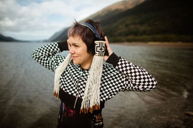 A woman wearing a black and white sweater and headphones with a black and white Indigenous design stands in front of water