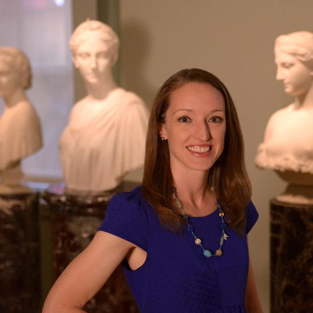 A photograph of a woman standing in an art gallery with busts behind her. 