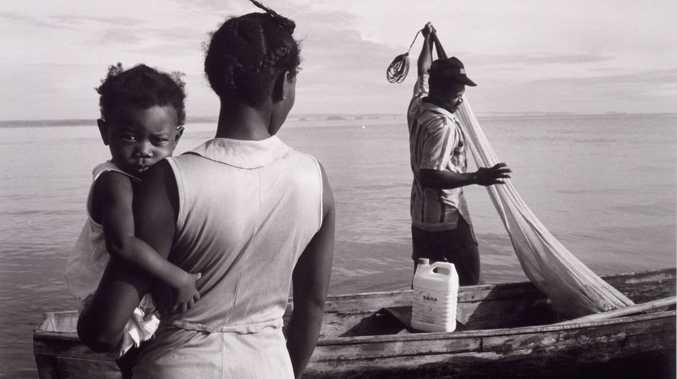 A photograph of a baby being held by a woman with a fisherman in the background