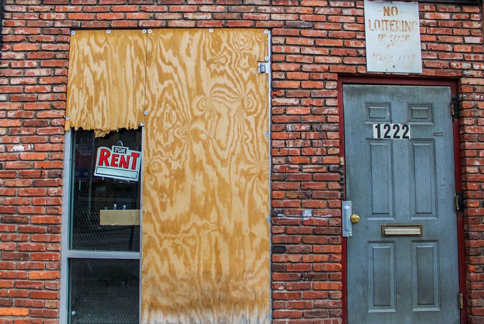 The exterior of a building with two doors, one boarded up with a "For Rent" sign to the left and one with a "No Loitering" sign above.