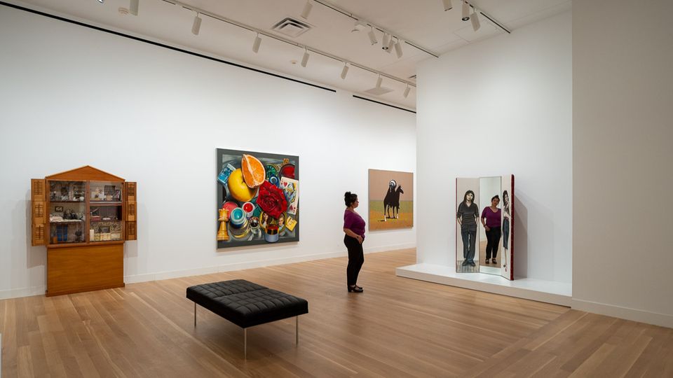 A person standing in a gallery is looking to a three-paneled artwork. The center panel is a mirror.