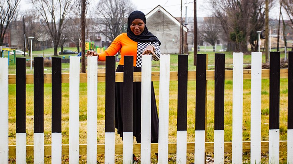 An African American girl wearing a black hijab looks directly at the camera, smiling. In front of her is a fence painted to resenble piano keys. Her fingers rest atop the fence.