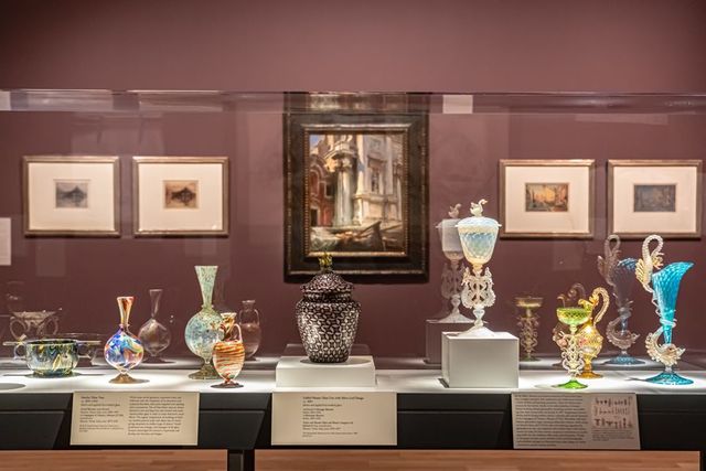 Exhibition gallery showing cases of glass artworks in the foreground and paintings against a dark red wall in the background.