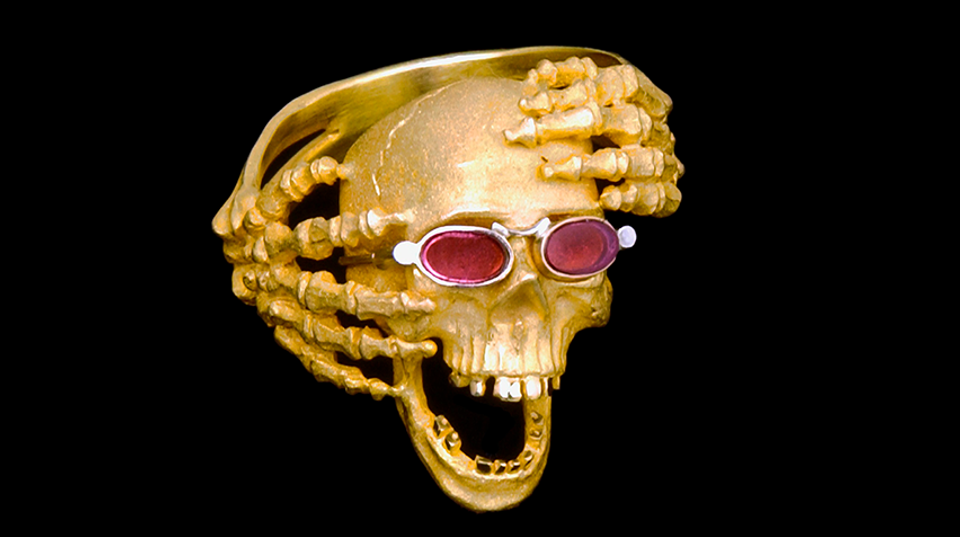 Sculpture that shows gold skeleton hands reaching around a gold skull that is wearing rose-colored glasses.
