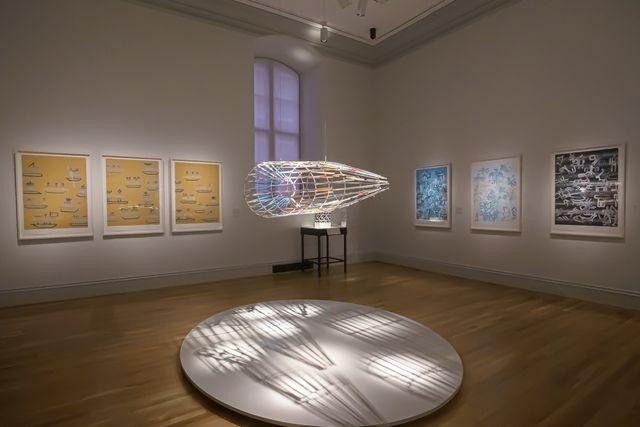 A wide angle shot of six prints on the wall, divided by a hanging glass cylindrical artwork