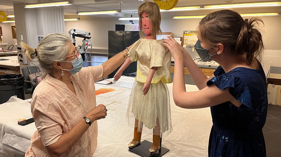 Two conservators, both wearing masks, re-dress the Gladys doll.