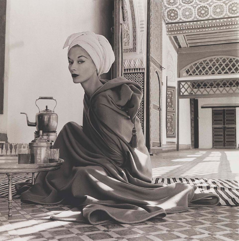 A photograph of a woman sitting in an interior with a tea pot and white scarf around her head.