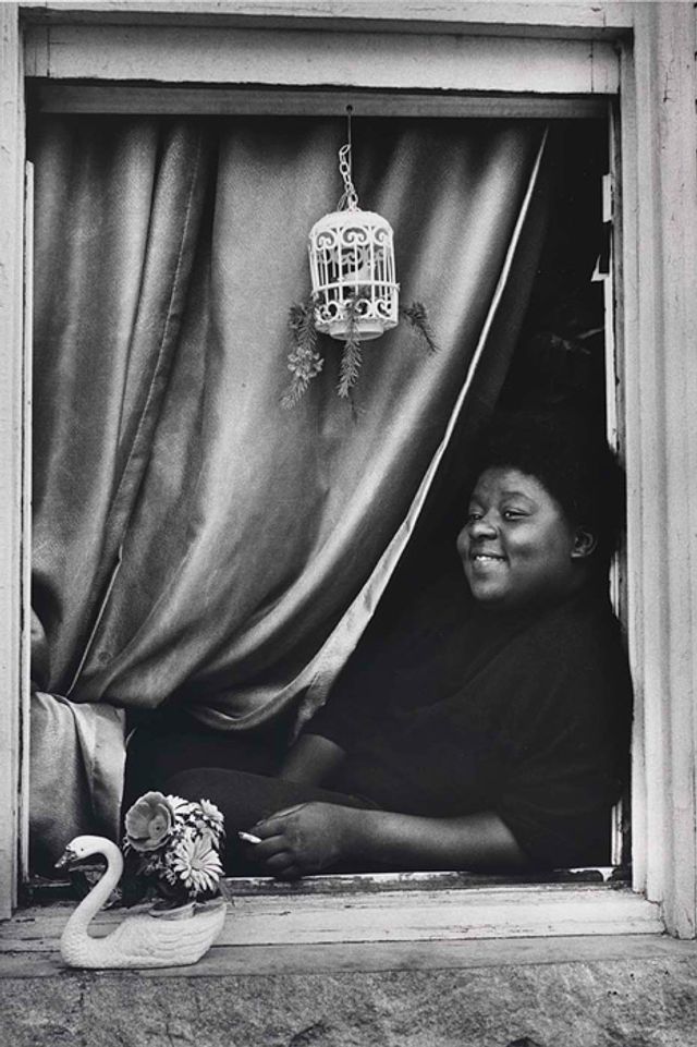 A gelatin silver print of a woman leaning in her window looking out with a curtain behind her. 