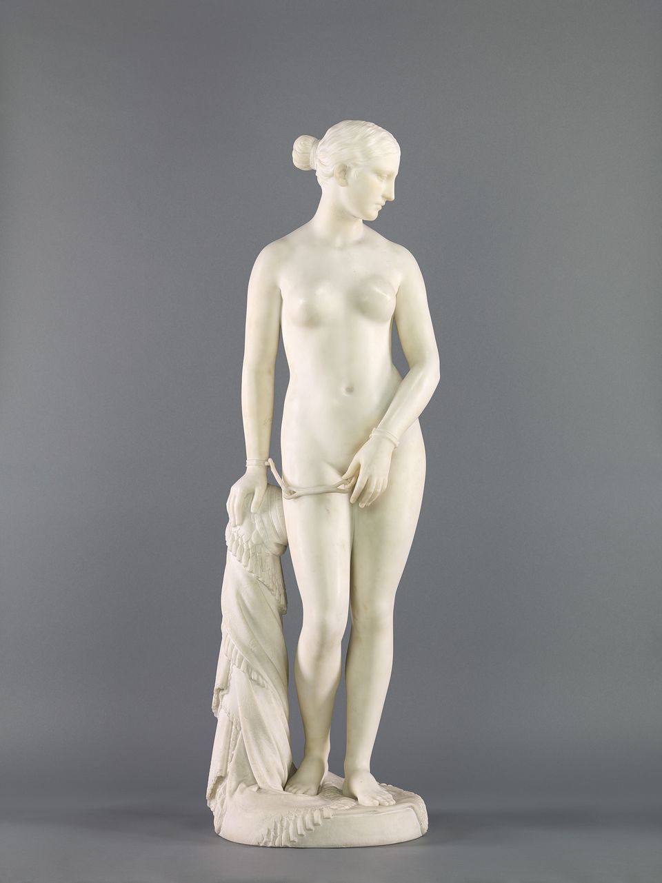 White marble sculpture depicting a nude woman standing with handcuffs on both wrists. A draped cloth is falling from one arm.