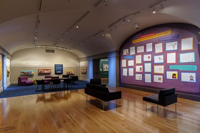 Museum galleries showing cards of reader responses hanging on a purple wall and a reading space with couches and rails of books on the far side of the room.