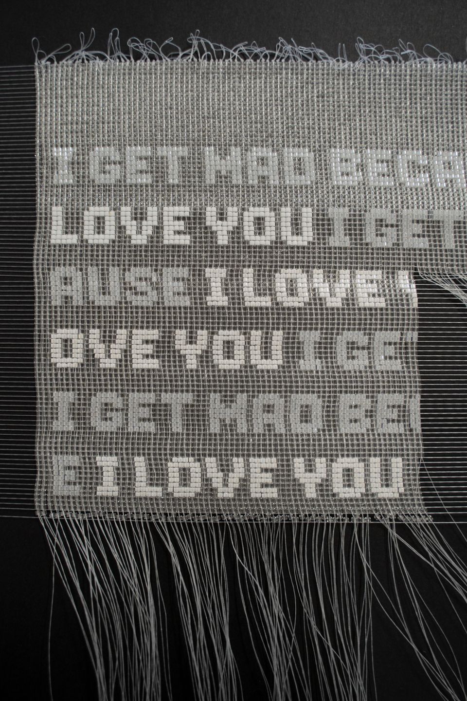 A detail of a woven artwork that is in progress. Text is written in beads. It repeats "I get mad because I love you."