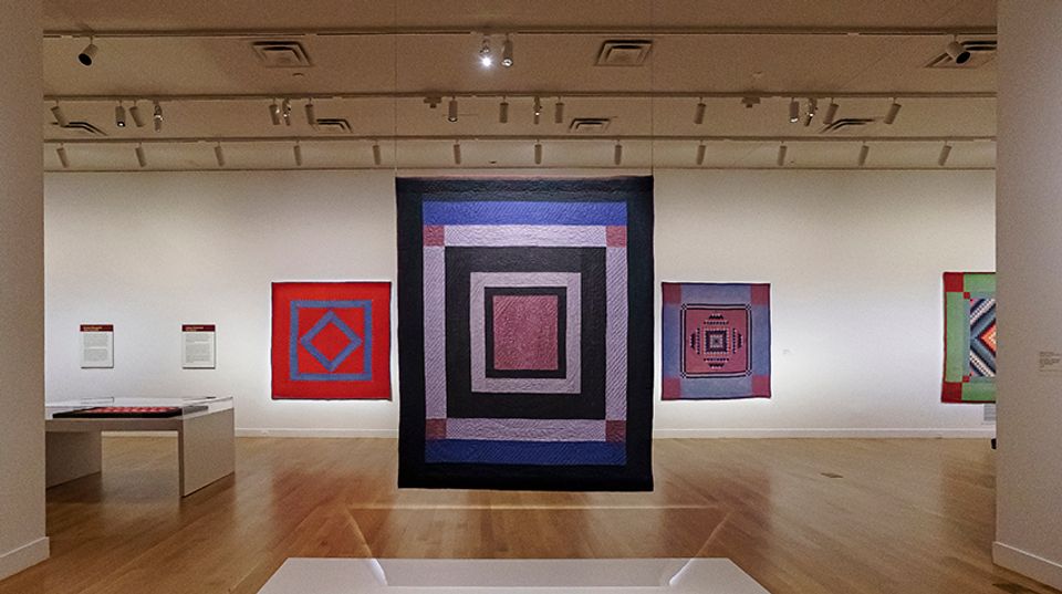 A quilt with large, concentric squares hang in a gallery. Quilts are mounted on the wall behind it.