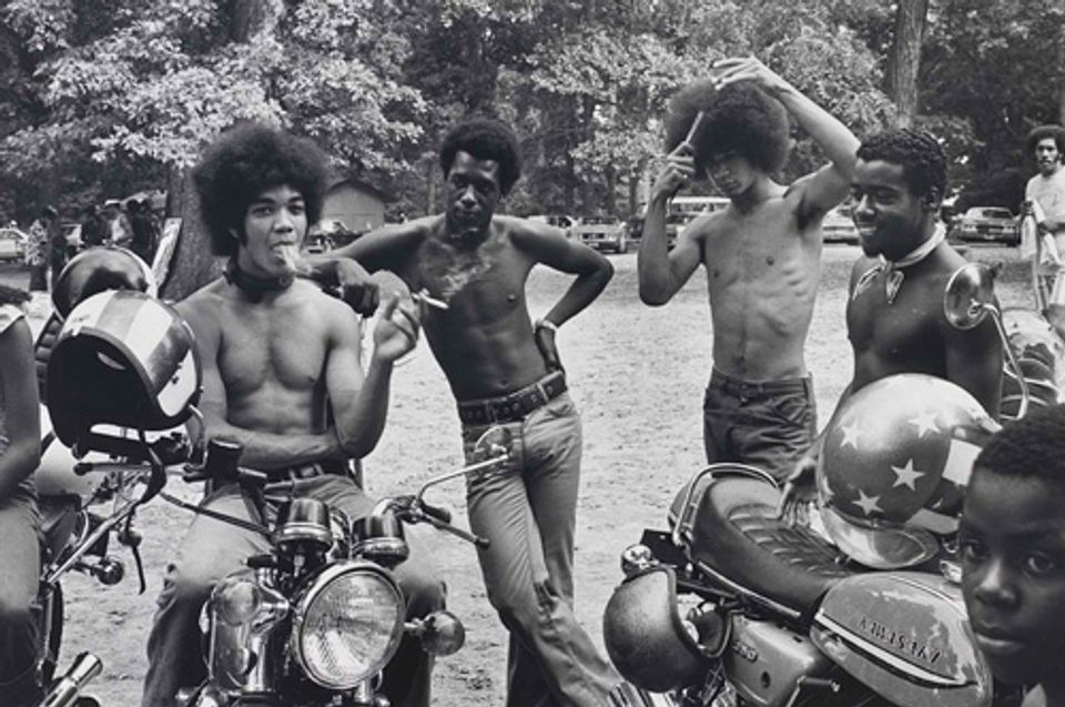 A gelatin silver print of four main male figures around motorcycles with trees in the background.
