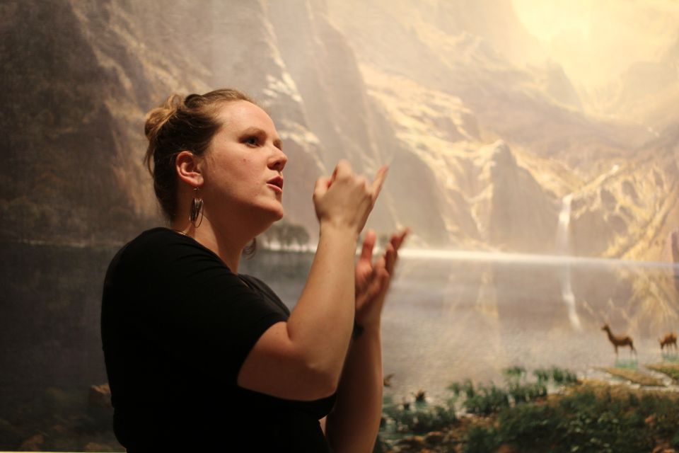 A photograph of a woman using her hands to sign in front of a landscape painting.