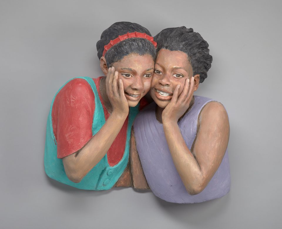 Painted bust depicting two young women. They are standing cheek to cheek, each with a hand to their face. 