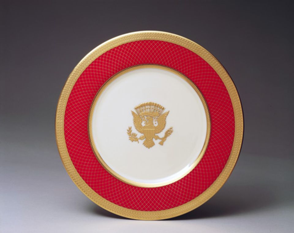 An image of a porcelain service plate with gold rim, red outer circle and crest. 