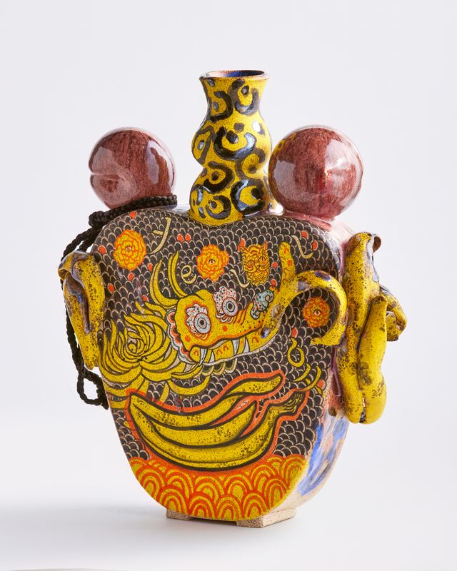 Colorful ceramic vase in yellows, reds, and blues, featuring an intricate textured dragon design flanked by two peeled bananas and two spherical handles on either side of a patterned neck, set against a nondescript background.