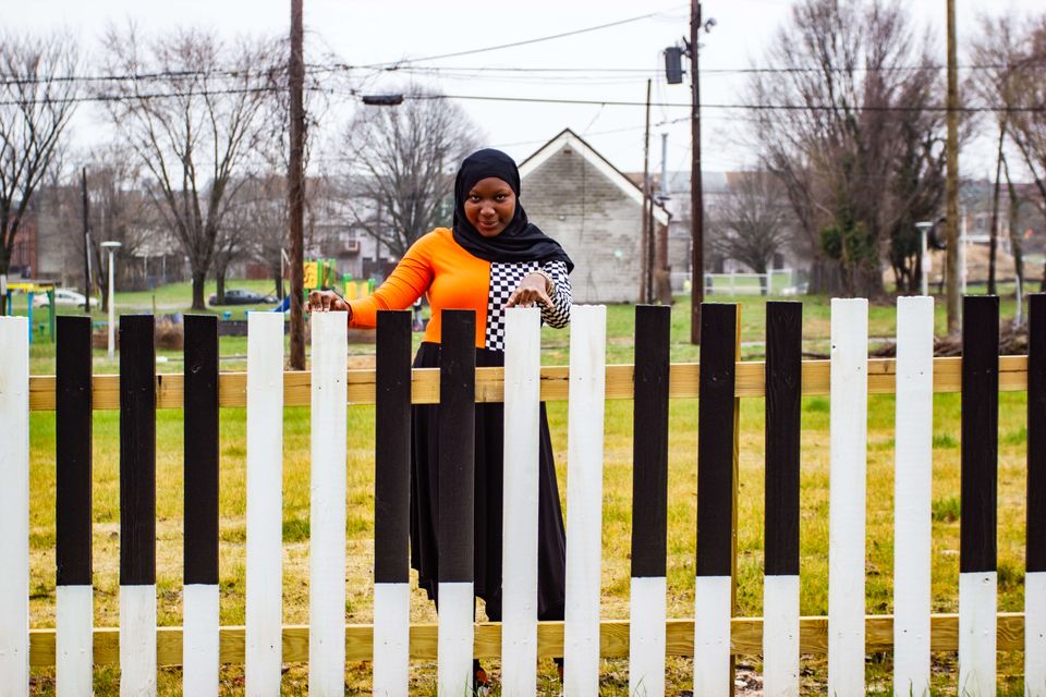 An African American girl wearing a black hijab looks directly at the camera, smiling. In front of her is a fence painted to resenble piano keys. Her fingers rest atop the fence.