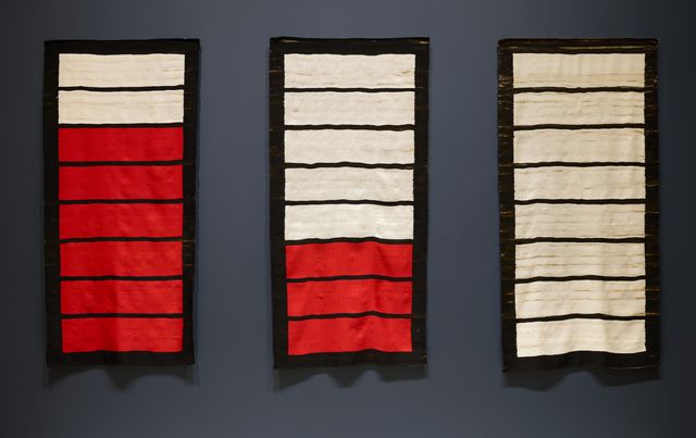 Three woven vertical rectangles hanging on a dark gray wall, each containing varying numbers of red and white segments.