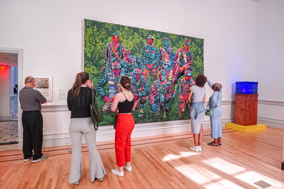 Groups of visitors stand in front of a large-scale portrait quilt which is hanging on a gallery wall.