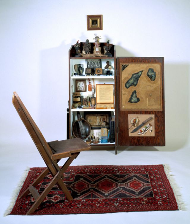 Stout's mixed media with a chair on a carpet and a shelf behind it.