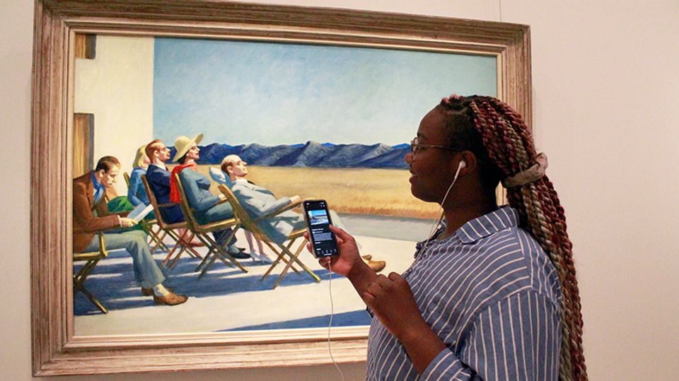 A photograph of a person standing in front of an artwork. She is wearing earbuds and holding her phone up to the work.