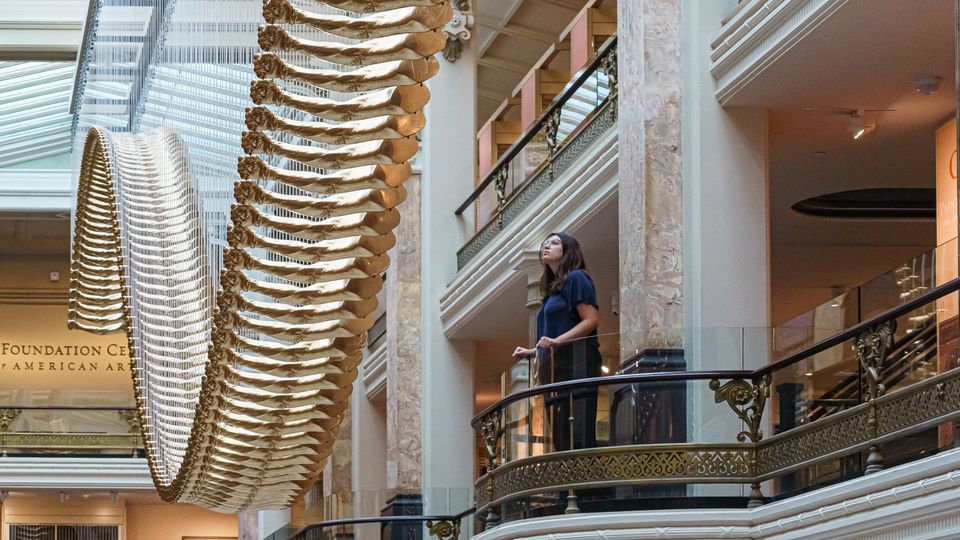 A person stands on a balcony and looks up at an artwork made of golden cast arms.