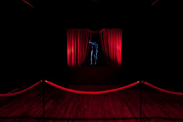 Installation image of a black room. There is a red curtain in the background and a red, velvet rope in the foreground. In between them is a blue, holographic image of a figure.