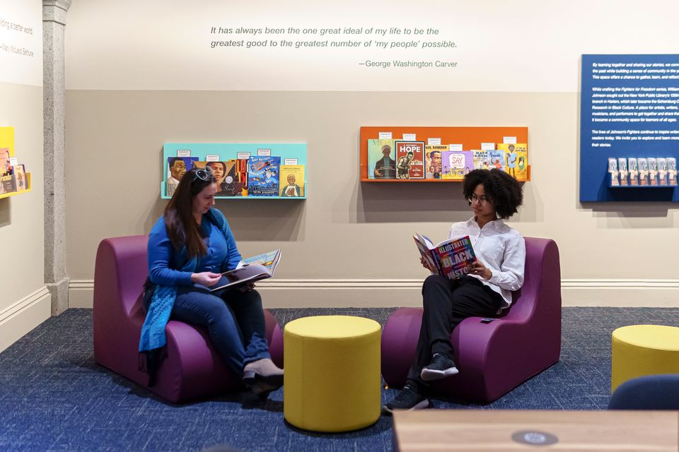 Two visitors sit on purple chairs reading books. There are rails of books on the wall behind them and round, yellow stools around them.
